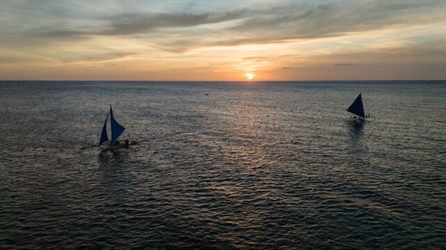 Aerial view of traditional Paraw sailboats sailing on the calm ocean at sunset in Boracay, Philippines. A beautiful tropical seascape during golden hour with vibrant orange sky