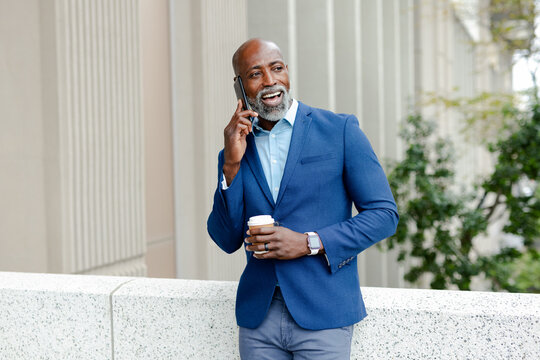 African american man leaning on railing at office, holding coffee, talking on smartphone