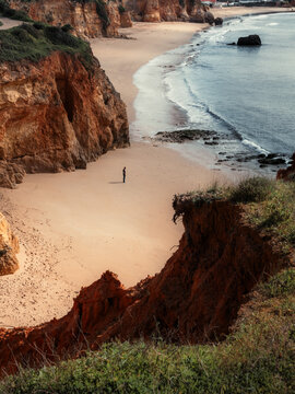 View of golden sands meeting the blue sea, framed by rugged, reddish cliffs under a soft sky, creating a tranquil coastal scene, Alvor, Faro District, Portugal.