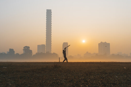 View of a lone cricketer silhouetted against the hazy dawn, with Kolkata's skyline piercing the golden mist, a tranquil moment frozen in time, Kolkata, West Bengal, India.
