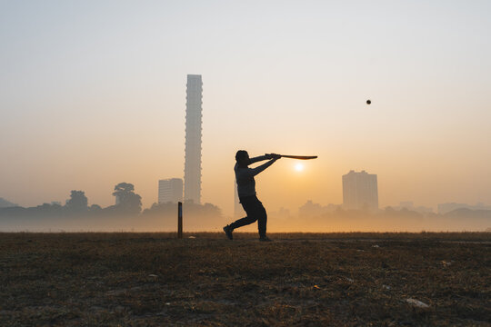 View of a lone cricketer silhouetted against the warm glow of the rising sun, playing a shot with cityscape in the misty background, Kolkata, West Bengal, India.
