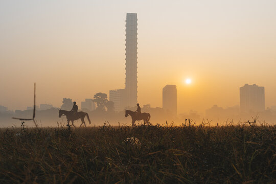 View of silhouetted riders astride horses traversing a sun-kissed field, with the iconic, towering 42 building piercing through the misty Kolkata skyline, Kolkata, West Bengal, India.