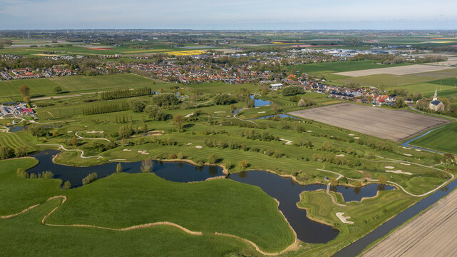 Aerial view of a golf course's green fairways and tranquil water hazards meet Winkel's charming village edge under a vast sky, Winkel, Noord-Holland, Netherlands.