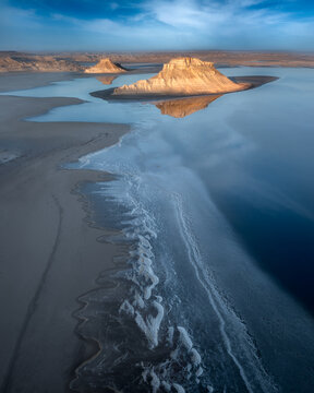 Aerial view of textured white salt formations meet the still, deep blue water reflecting the desert buttes under a clear sky, Aktau, Mangystau Region, Kazakhstan.