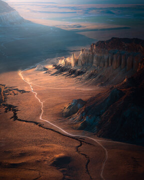 Aerial view of sunlit cliffs casting long shadows across a rugged, arid landscape, the winding path contrasting against the earth, Aktau, Mangystau Region, Kazakhstan.