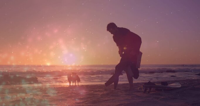 Surfer pulling wetsuit off after surfing on sunset beach, lens flare showing surfboard and dogs
