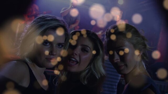 Three women leaning for concert selfie, raising smartphone, singing with bokeh drifting over faces