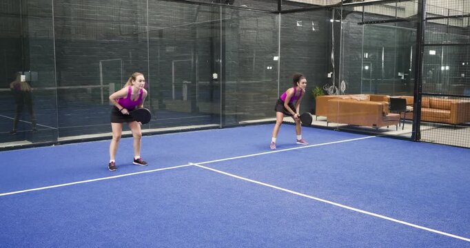 Diverse female friends entering padel court in sportswear, rallying ball with rackets to warm up