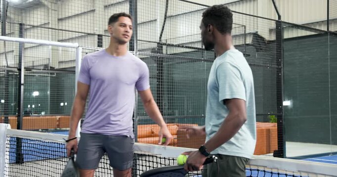 Diverse male friends entering padel court walking to net bouncing balls with rackets warming up