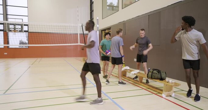 Diverse male teammates placing duffel on bench picking volleyball moving to net practicing passing