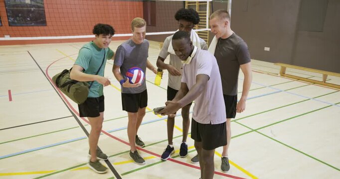 Diverse male team gathering as newcomer arriving with ball, angling phone for selfie on gym court