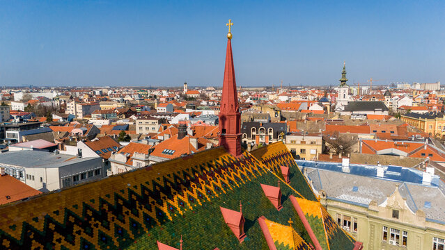 Aerial view of the intricate, patterned roof tiles lead to a striking red steeple against a backdrop of buildings stretching to the horizon, Sad, Vojvodina, Serbia.