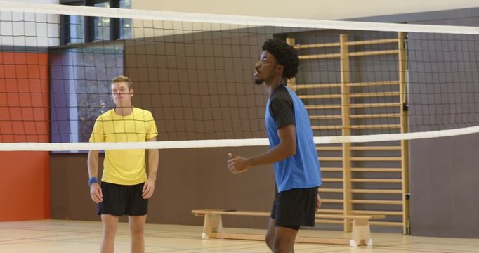 Diverse male players in jerseys reacting to serve in gym, blue player contacting ball above net