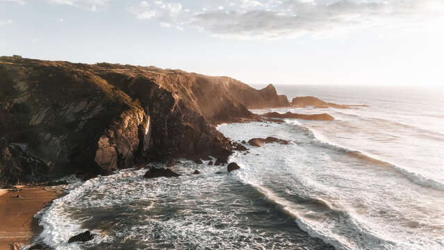 Aerial view of rugged cliffs meet the churning Atlantic, where golden light paints the crashing waves and rocky outcrops, Cabe&Atilde;&sect;o da Arv&Atilde;&copy;loa, Beja, Portugal.