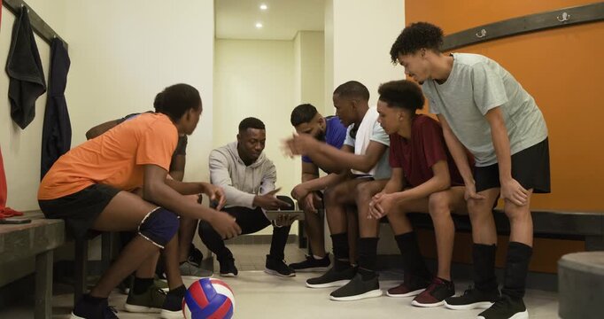African-American coach kneeling pointing to clipboard in locker room urging men to study play