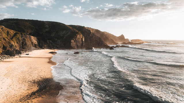 Aerial view of golden sands meeting the frothy, white-tipped waves under the warm glow of the setting sun, framed by rugged cliffs, Cabeco da Arveloa, Portugal.