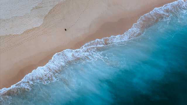 Aerial view of a lone figure strolls along the sandy shore as azure waves crash, creating a stunning contrast of color and texture, Alvor, Faro, Portugal.