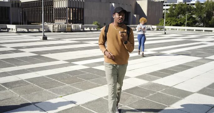 African American man walking across tiled plaza holding phone to mouth, taking call adjusting cap