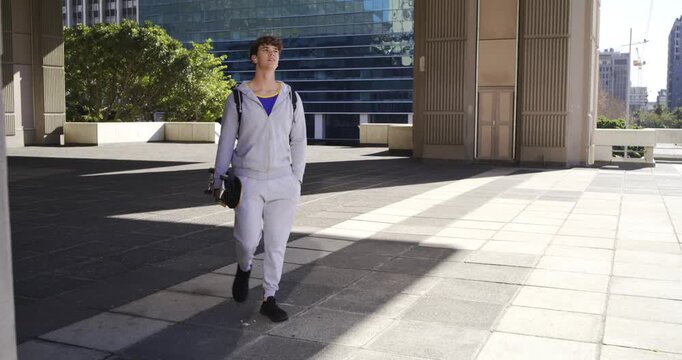Man walking forward from shaded plaza into bright sunlight, carrying skateboard and backpack