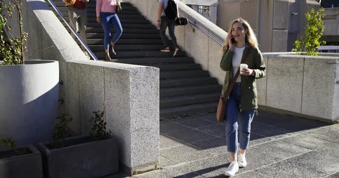 Blonde woman continuing smartphone call, wearing parka and holding coffee while walking down stairs