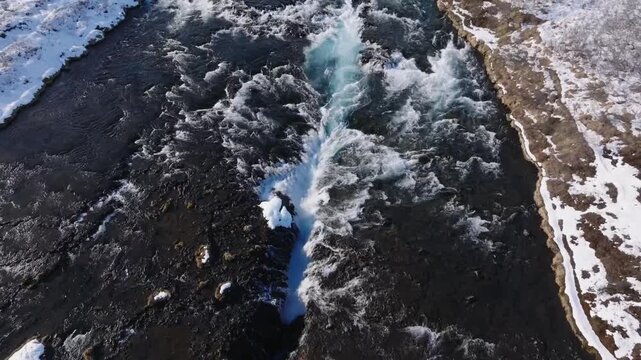 Stunning drone footage captures the vivid blue waters of Bruarfoss waterfall cascading through dramatic black volcanic rock formations surrounded by pristine winter snow in Iceland's wilderness.