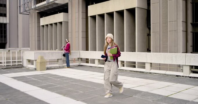 Adult female seeing phone, pushing off railing, walking on rooftop with backpack and notes to class