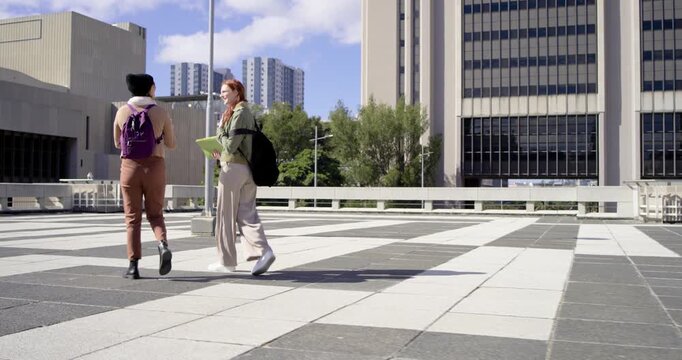 Two women talking and walking across tiled plaza, gesturing with green folder; passerby phoning