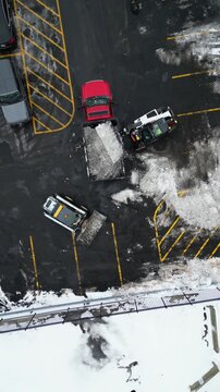 An overhead drone shot captures a skid steer loader depositing snow into the back of a red work truck in an asphalt parking lot during winter maintenance.