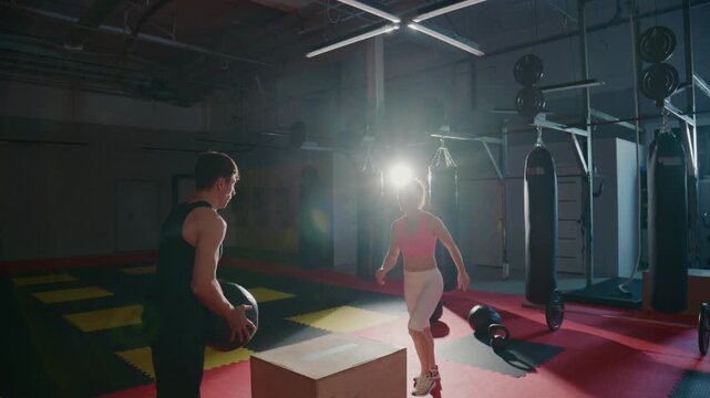 Young woman trains with coach in a gym. Coach guides her movements with focused attention. Gym lights cast bright shadows across the floor. Energy and discipline fill the training space