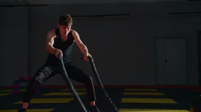 Athlete grips rope with intense focus and power. Dark gym lights highlight sweat and effort. Red mat contrasts with yellow safety stripes. Body moves low, strong, and controlled