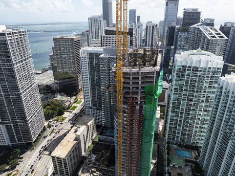 Aerial view of a skyscraper under construction piercing the skyline amidst the bustling cityscape of Brickell, its crane a stark contrast against the azure bay, Miami, Florida, United States.