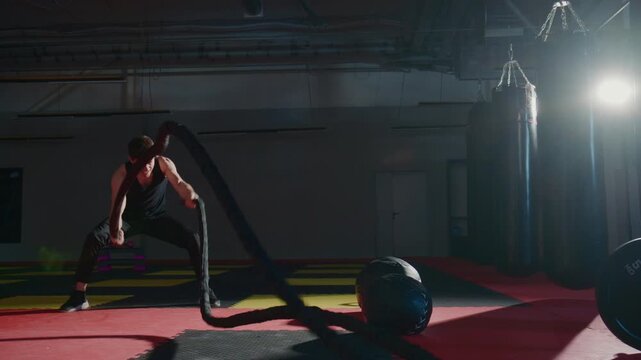 Athletic man grips heavy battle rope with intense focus. He performs explosive, powerful movements in dim, industrial gym. Bright spotlight enhances muscle definition and effort