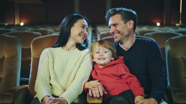 Asian mother white father smiling child, cozy theater seats, toddler in red hoodie cuddling between parents, parents exchange warm glance, dim ambient lights, candid family moment capturing joy