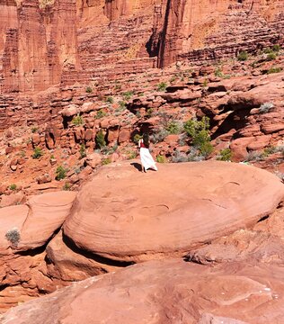 Aerial view of a solitary figure in a white dress stands atop a rounded, red rock formation, dwarfed by the towering, layered cliffs of the Moab desert, Utah, United States.