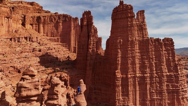 Aerial view of a lone climber ascends a towering sandstone pillar amidst the sun-baked, rugged landscape, the Fisher Towers casting long shadows under a vast sky, Moab, Utah, United States.