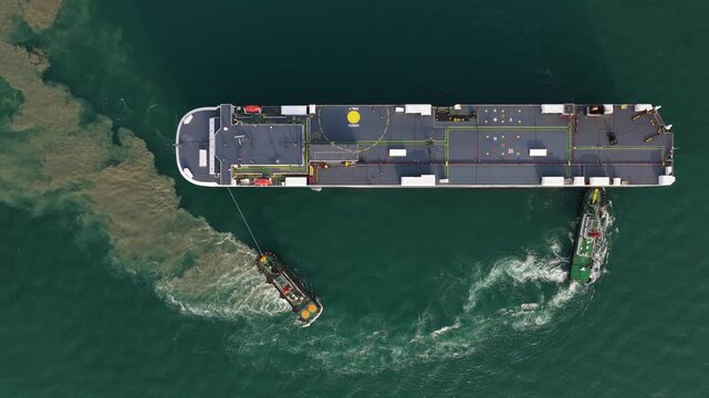 Aerial top down view of a huge ro ro cargo ship, a vehicle carrier, sailing on calm green water. Tugboats assist the large vessel, with exhaust smoke billowing from the funnel