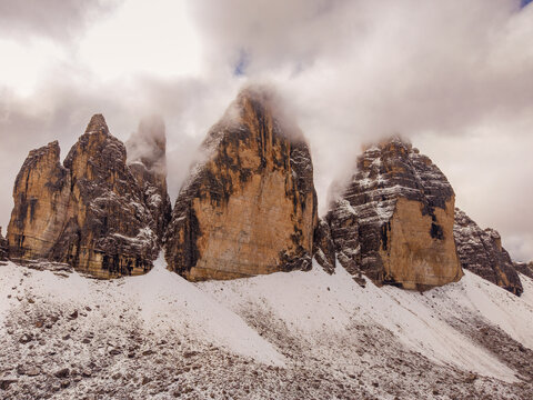 Aerial view of jagged peaks of Tre Cime di Lavaredo piercing through swirling clouds and dusted with snow, Tre Cime di Lavaredo, Veneto, Italy.