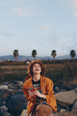 Happy woman with curly hair wearing orange jacket laughs holding smartphone outdoors near rocks with palm trees and mountains in background during casual lifestyle moment.