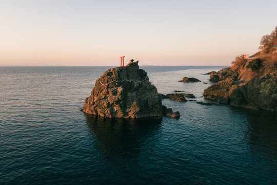Aerial view of Tsunakakeiwa Rock Shrine, a rugged island adorned with a vibrant red torii gate under the fading light, Ehime, Shikoku, Japan.