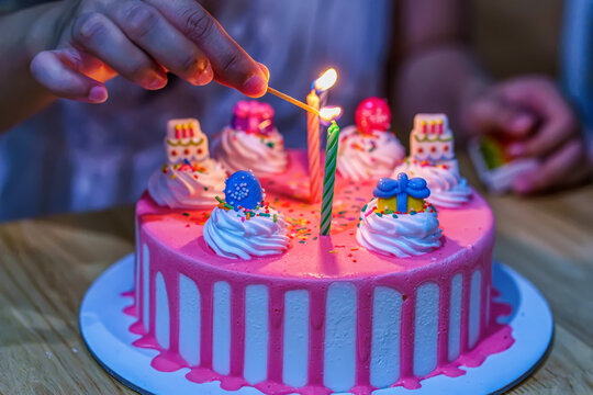 A hand lighting candles on a pink birthday cake decorated with cream and colorful toppings.