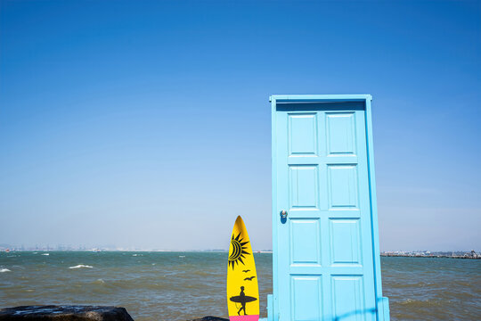 Minimal seascape with a bright blue door and yellow surfboard standing on coastal rocks against a wide blue sky and choppy sea, creating a whimsical travel scene.
