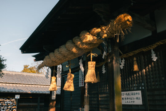 View of a thick, woven rope hanging from a traditional Japanese shrine entrance, its golden hues contrasting with the dark wood, Kanonji, Kagawa, Japan.