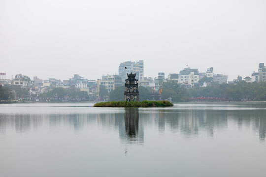 Hanoi, Vietnam - 18 January 2026: View of the Turtle Tower rising from the serene waters of Hoan Kiem Lake, its reflection shimmering amidst the city's hazy skyline.