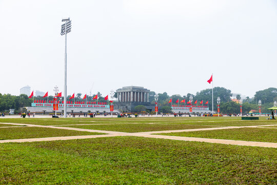 Hanoi, Vietnam - 18 January 2026: View of the Ho Chi Minh Mausoleum, a solemn grey structure framed by vibrant red flags, set against a sprawling green field.