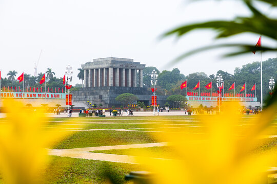 Hanoi, Vietnam - 18 January 2026: View of the imposing Ho Chi Minh Mausoleum, framed by blurred yellow foliage and vibrant red flags fluttering proudly against a muted sky.