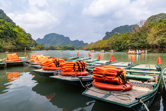 View of vibrant orange life jackets stacked on boats awaiting passengers amidst lush green hills and serene waters, Trang An, Ninh Binh, Vietnam.