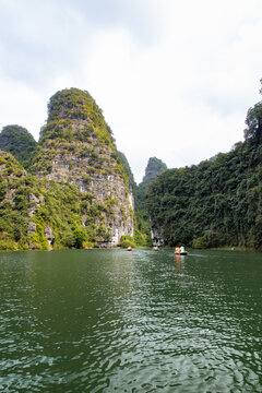 View of a serene river reflecting towering, verdant cliffs where boats glide peacefully under a soft sky, Trang An, Ninh Binh, Vietnam.