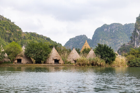 View of rustic huts with conical thatched roofs line the tranquil waters reflecting the surrounding lush greenery and towering limestone karsts, Trang An, Vietnam.