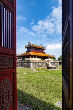 View of ornate red doors frame a golden-roofed pavilion on a green lawn under a bright blue sky, Hue Imperial City, Hue, Vietnam.