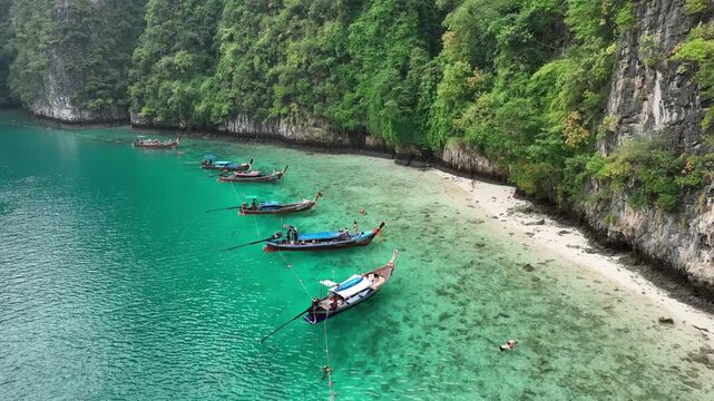 Aerial view of traditional longtail boats contrasting against the turquoise waters meeting the sandy beach and lush green cliffs, Kho Phi Phi Island, Thailand.